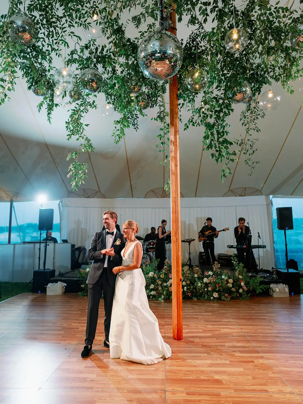 Evening interior Sperry sailcloth tent with center pole and greenery decor. Bride and groom on dance floor. Band stage with band